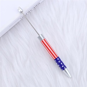 May include: A pen with a patriotic design featuring red and white stripes and blue stars. The pen has a silver tip and a long, thin metal rod extending from the top.