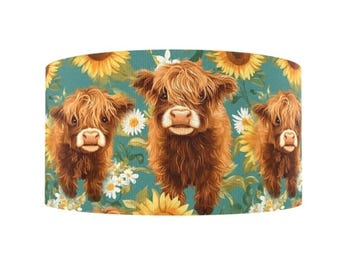 May include: A cylindrical lampshade featuring three Highland cows with shaggy brown fur. The background is a teal color with yellow sunflowers and white daisies. The lampshade is a decorative home decor item.