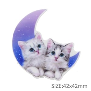 May include: Two white kittens with blue eyes are nestled together on a purple crescent moon with a starry background. The moon is 42 mm wide and 42 mm tall.