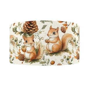 May include: A cylindrical lampshade with a watercolour design of two squirrels holding acorns, surrounded by pine cones, leaves, and berries. The colour palette includes shades of brown, orange, and green on a cream background. A decorative home decor item.