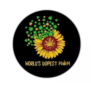 May include: A black circle with a yellow sunflower with green marijuana leaves in the center. The sunflower is surrounded by green marijuana leaves. The text "WORLD'S DOPEST MOM" is written below the sunflower.