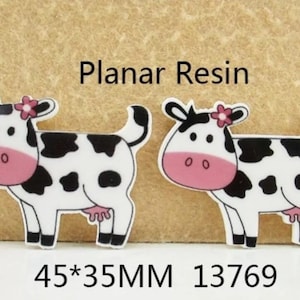 Pode incluir: Dois enfeites de resina de vaca preta e branca com detalhes em rosa e uma flor na cabeça. As vacas medem 45 mm por 35 mm e o número do produto é 13769.