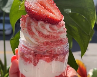 May include: A clear glass jar filled with layers of pink cake, white frosting, and crushed pink candy. A strawberry slice sits on top of the frosting.