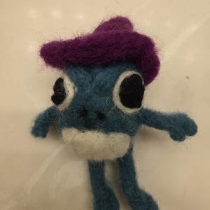 May include: A blue felt frog wearing a purple felt hat. The frog has white felt for its belly and black felt for its eyes.