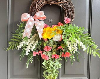 May include: A spring-themed wreath with a pink and white gingham bow, yellow flowers, and green foliage. The wreath is hanging on a dark gray door.