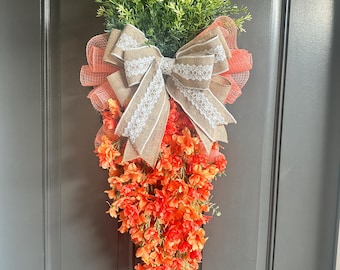 May include: A carrot-shaped Easter door wreath with a burlap bow and orange flowers. The wreath is decorated with a white lace ribbon and green foliage.