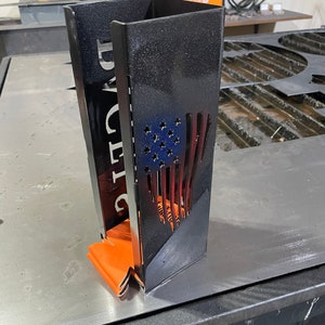 May include: A black metal fire pit with a cut-out American flag design and the word "DADDY" on the side. The fire pit is sitting on a metal surface.