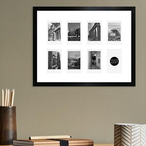 May include: A black picture frame with a white mount and nine black and white photographs of various locations. The frame is hanging on a light brown wall above a wooden desk with a wicker chair, a lamp, and a mug.