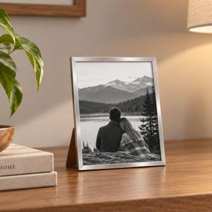 May include: A rectangular silver metal picture frame containing a black and white photograph of two people sitting together outdoors, with a mountain and lake landscape in the background, displayed on a wooden surface.