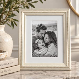 May include: A rectangular cream-colored picture frame with a beaded inner border detail. The frame holds a black and white photograph of a smiling family consisting of two adults and a child, all captured in a close-up, joyful embrace.