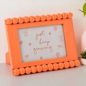 May include: An orange picture frame with a beaded edge. The frame displays a white background with the words "just keep growing" in cursive, and small flower and bow illustrations. The frame is supported by a stand.
