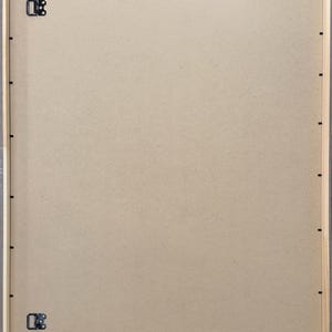 May include: Back of a framed artwork, showing the wooden frame, cardboard backing, and hanging hardware. The frame is light-colored wood with a gray border. Metal clips and hooks are attached for hanging.