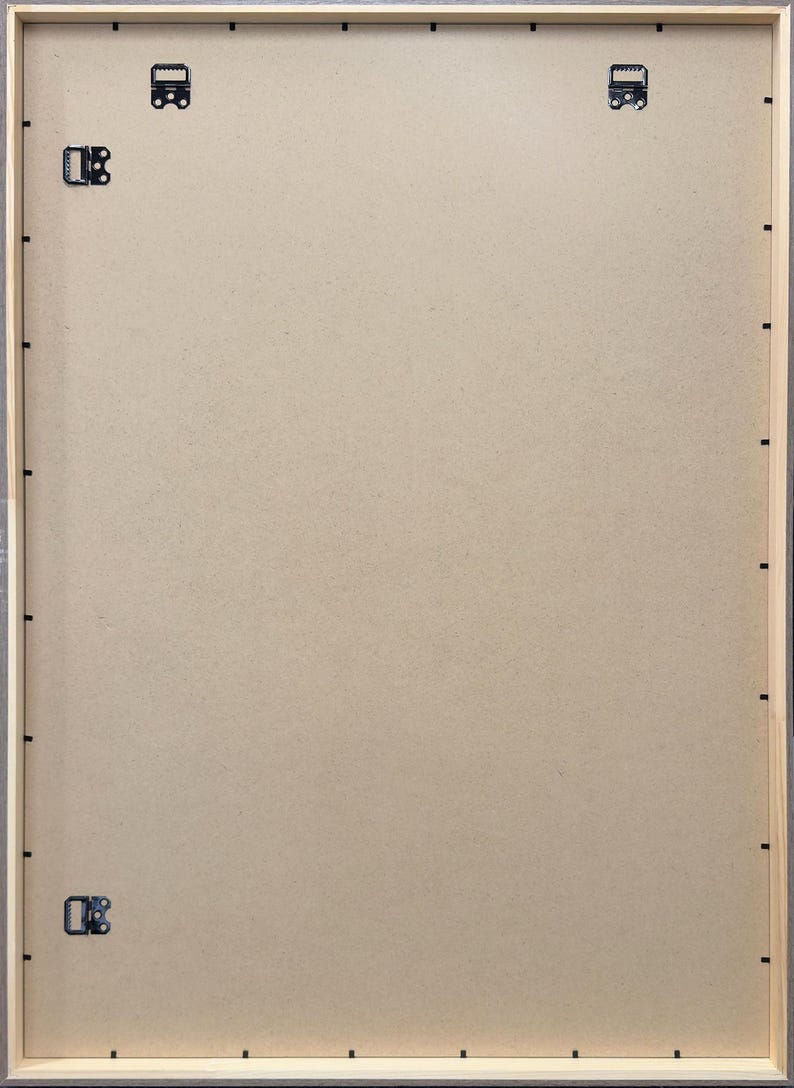 May include: Back of a framed artwork, showing the wooden frame, cardboard backing, and hanging hardware. The frame is light-colored wood with a gray border. Metal clips and hooks are attached for hanging.