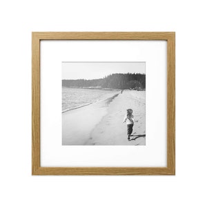 May include: A black and white photograph of a child running on a beach. The child is wearing a light coloured shirt and dark trousers. The beach is empty except for the child and two other people in the distance.