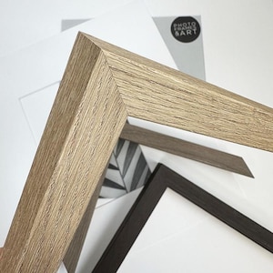 May include: Close-up of a light brown wooden picture frame corner, showcasing the wood grain texture. Other frames in dark brown and gray are visible, along with a black and white leaf drawing and a white mat. The text "PHOTO FRAMES & ART" is visible.