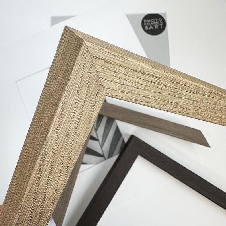 May include: Close-up of a light brown wooden picture frame corner, showcasing the wood grain texture. Other frames in dark brown and gray are visible, along with a black and white leaf drawing and a white mat. The text "PHOTO FRAMES & ART" is visible.