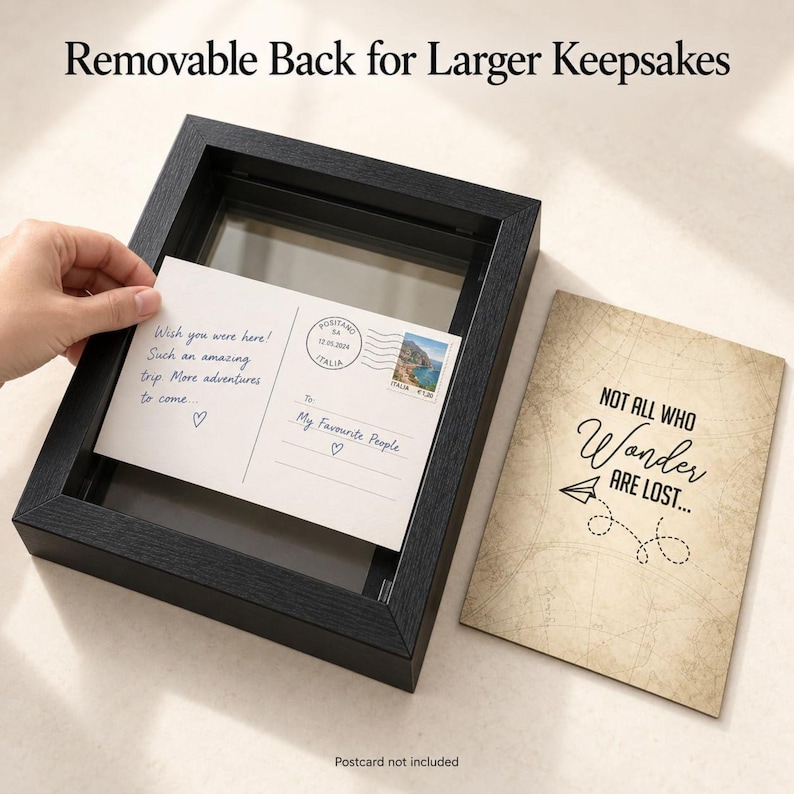 May include: A black shadow box frame with a removable back for larger keepsakes. A postcard with the text "Wish you were here!" is being placed inside. Next to it is a tan card with the text "NOT ALL WHO WONDER ARE LOST..." and a paper airplane.