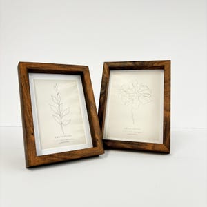 May include: Two wooden photo frames with a warm brown finish. Each frame holds a print with a delicate line drawing of a plant. The frames are angled slightly, showcasing the artwork within. The frames are 15.24 cm x 20.32 cm.