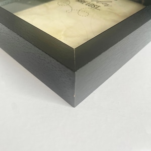 May include: A black wooden shadow box frame containing a cream-colored paper insert. The visible text on the paper reads "ARE LOST..." in a dark font, accompanied by a small dotted line graphic. The frame has a smooth, matte finish.
