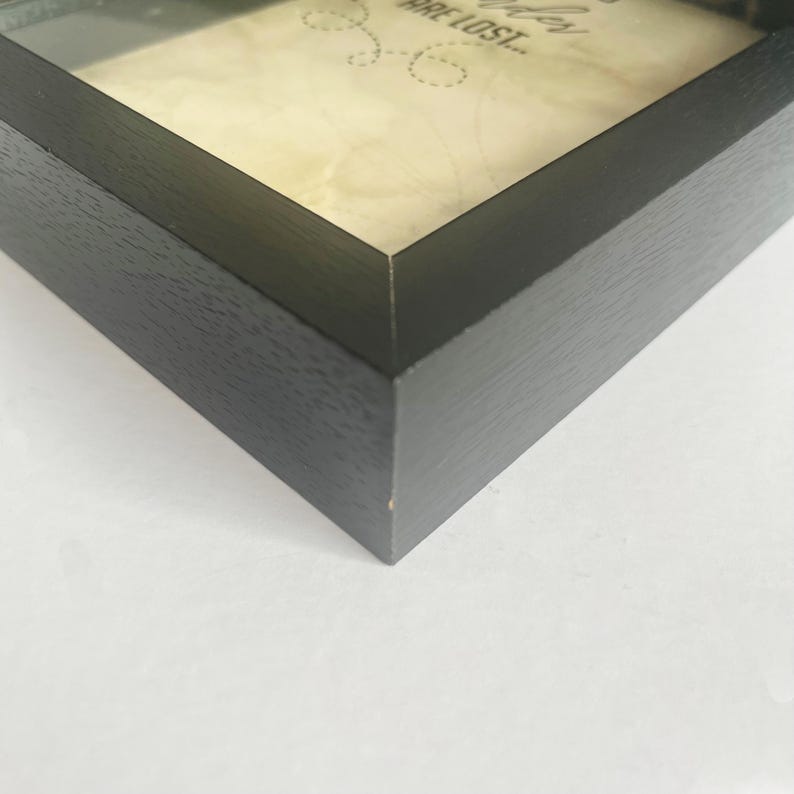 May include: A black wooden shadow box frame containing a cream-colored paper insert. The visible text on the paper reads "ARE LOST..." in a dark font, accompanied by a small dotted line graphic. The frame has a smooth, matte finish.