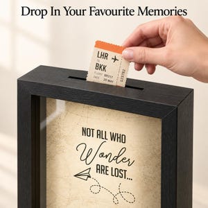 May include: A black shadow box frame with a slot at the top for inserting items. The interior features a beige background with a map pattern and the text "NOT ALL WHO Wonder ARE LOST..." with a paper airplane graphic. Text above reads "Drop In Your Favourite Memories".