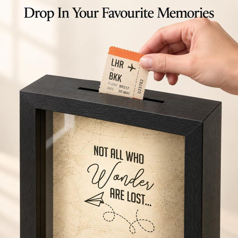 May include: A black shadow box frame with a slot at the top for inserting items. The interior features a beige background with a map pattern and the text "NOT ALL WHO Wonder ARE LOST..." with a paper airplane graphic. Text above reads "Drop In Your Favourite Memories".
