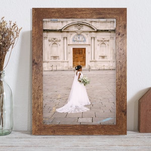 May include: A framed photograph of a bride wearing a white wedding dress and veil, standing in front of a building with a large doorway. The bride is holding a bouquet of flowers.
