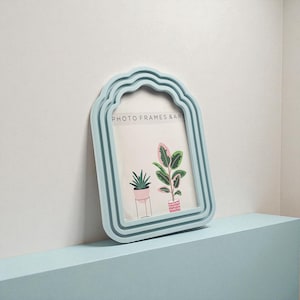 May include: A pale blue photo frame with a decorative, arched top. The frame holds a print of two potted plants. The text "PHOTO FRAMES & ART" is printed on the print. The frame is resting on a pale blue surface.