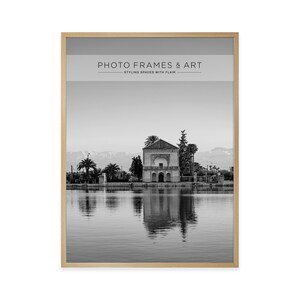 May include: Black and white framed print featuring a serene landscape with a building reflected in calm water. The print is in a light wood frame. Text at the top reads "PHOTO FRAMES & ART" and "STYLING SPACES WITH FLAIR".