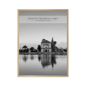 May include: Black and white framed print featuring a serene landscape with a building reflected in the water. The print is in a light wood frame. The text at the top reads "PHOTO FRAMES & ART" and "STYLING SPACES WITH FLAIR".