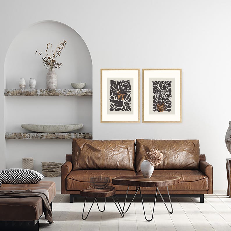 May include: Two framed abstract art prints with botanical designs in black and tan hues. The artwork hangs above a brown leather sofa, complemented by a wooden coffee table and a matching ottoman. The room has a neutral color palette.