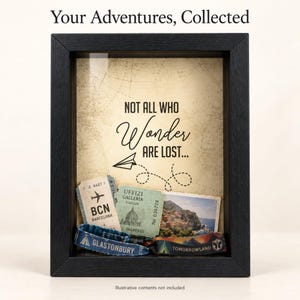 May include: A black shadow box frame containing a beige background with the text "Your Adventures, Collected" and "NOT ALL WHO Wonder ARE LOST..." with a paper airplane graphic. Inside are various travel tickets and two fabric wristbands.