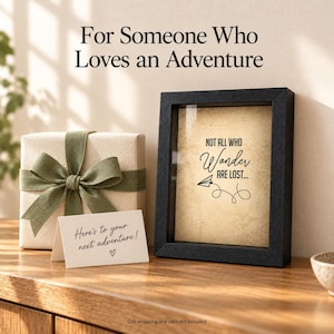May include: A black shadow box frame containing a beige paper insert with the text "NOT ALL WHO Wander ARE LOST..." and a paper airplane graphic. Beside it sits a gift box wrapped in cream paper with a green ribbon and a small white note card.