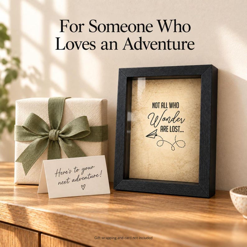 May include: A black shadow box frame containing a beige paper insert with the text "NOT ALL WHO Wander ARE LOST..." and a paper airplane graphic. Beside it sits a gift box wrapped in cream paper with a green ribbon and a small white note card.