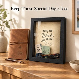 May include: A black shadow box frame containing a beige background with the text "NOT ALL WHO WONDER ARE LOST..." and a paper airplane graphic. Inside the frame are two travel tickets and a wristband. Next to it is a brown leather journal.