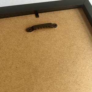 May include: The back of a black picture frame featuring a brown fiberboard backing. A metallic sawtooth hanger is securely attached to the top center of the frame, designed for wall mounting. The frame edges are smooth and dark in color.
