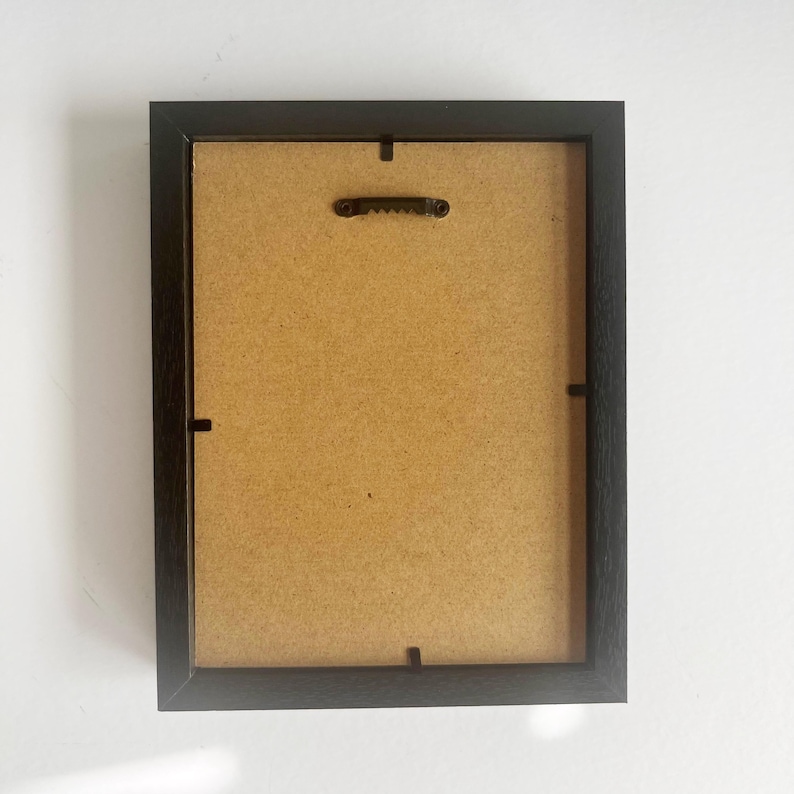 May include: A rectangular picture frame shown from the back, featuring a dark brown wooden border, a light tan fiberboard backing, and a central black metal sawtooth hanger attached to the top edge for wall mounting purposes.