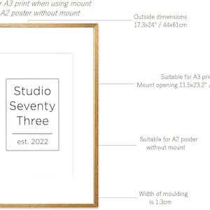 May include: A gold-colored picture frame with a white mat. The frame is 17.3 inches by 24 inches and has a 1.3 inch wide molding. The frame is suitable for an A3 print with a mount or an A2 poster without a mount. The mount opening is 11.5 inches by 23.2 inches. The frame has a logo that says "Studio Seventy Three est. 2022".