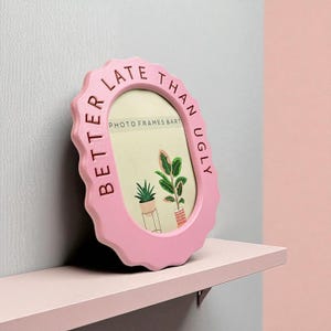May include: A pink, scalloped-edge photo frame with the text "BETTER LATE THAN UGLY" around the oval aperture. The frame displays a picture of two potted plants and the text "PHOTO FRAMES & ART".