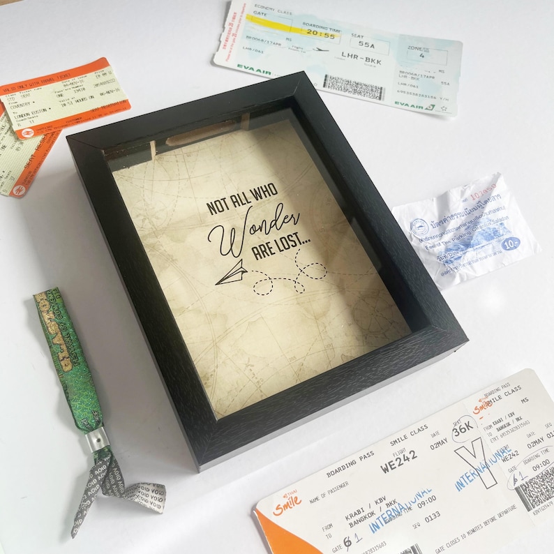 May include: A black shadow box frame containing a cream-colored background with a map print and the text "NOT ALL WHO Wonder ARE LOST..." accompanied by a paper airplane graphic. The frame is surrounded by various airline boarding passes.