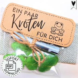 Puede incluir: Un pequeño paquete de regalo con caramelos verdes, atado con hilo. Una etiqueta marrón con el texto "EIN PAAR Kröten FÜR DICH" y una ilustración de rana de dibujos animados está adjunta. También es visible un pequeño icono de descarga PDF.