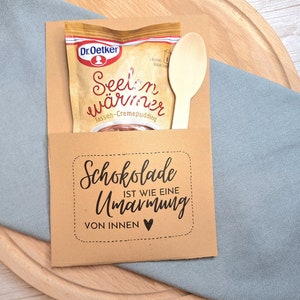 May include: A brown paper envelope with a small wooden spoon and a packet of chocolate pudding mix. The envelope has a black and white text that reads "Schokolade ist wie eine Umarmung von innen" with a small heart.