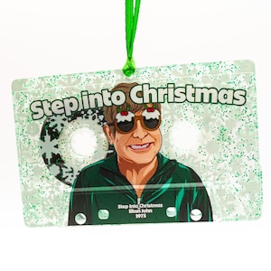 Könnte beinhalten: Ein klarer Weihnachtsschmuck in Form einer Kassette, bedeckt mit Glitzer. Es zeigt eine Cartoon-Illustration von Elton John mit ausgefallenen Sonnenbrillen. Der Text "Step into Christmas" steht über der Illustration, darunter "Elton John 1975".