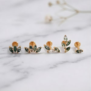 May include: A selection of gold-coloured stud earrings. Some earrings feature green and white stone accents in a leaf-like design, while others have clear, sparkling stones. The earrings are displayed on a white marble surface.