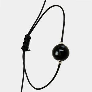 Peut inclure: Un bracelet en cordon noir doté d'une unique perle sphérique noire brillante, placée entre deux petites entretoises en métal argenté. Le bracelet est muni d'un fermoir coulissant réglable fabriqué dans le même cordon noir.