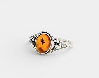 Pode incluir: Um anel de prata com uma pedra ovalada de cor laranja. O anel apresenta um design ornamental de filigrana em torno da pedra e na banda. A pedra apresenta um padrão único, adicionando à aparência distintiva do anel. Em fundo branco.