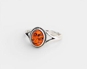 Pode incluir: Um anel de prata com uma pedra ovalada de cor âmbar. A pedra é engastada em uma moldura decorativa com uma borda em forma de corda. O anel tem uma banda simples com um design triangular em cada lado da pedra.