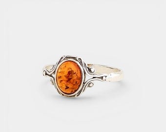Pode incluir: Um anel de prata com uma pedra de âmbar laranja translúcida de formato oval no centro. A banda de metal apresenta padrões decorativos intrincados e em espiral que emolduram a pedra, criando uma aparência detalhada e ornamentada.