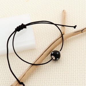 Peut inclure: Un bracelet noir réglable avec une seule perle ronde noire à pois blancs. Le bracelet est fait d'un cordon noir fin et est présenté sur une surface de couleur claire avec un élément en bois.
