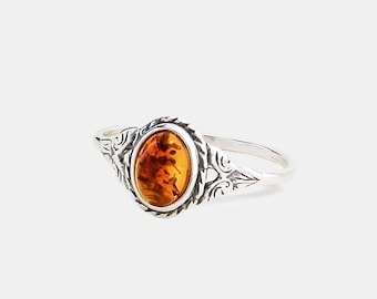 Pode incluir: Um anel de prata com uma pedra de âmbar oval e translúcida engastada num bisel decorativo. A banda e o engaste são trabalhados com padrões intrincados de volutas em relevo que se estendem ao longo dos lados do anel.